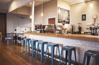 A cozy coffee shop interior featuring a wooden counter with a row of blue metal stools. A barista is preparing drinks behind the counter, surrounded by various coffee machines and accessories. Hanging light fixtures provide a warm ambiance, and a chalkboard menu is visible on the wall.