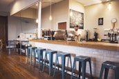 A cozy coffee shop interior featuring a wooden counter with a row of blue metal stools. A barista is preparing drinks behind the counter, surrounded by various coffee machines and accessories. Hanging light fixtures provide a warm ambiance, and a chalkboard menu is visible on the wall.
