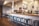 A cozy coffee shop interior featuring a wooden counter with a row of blue metal stools. A barista is preparing drinks behind the counter, surrounded by various coffee machines and accessories. Hanging light fixtures provide a warm ambiance, and a chalkboard menu is visible on the wall.