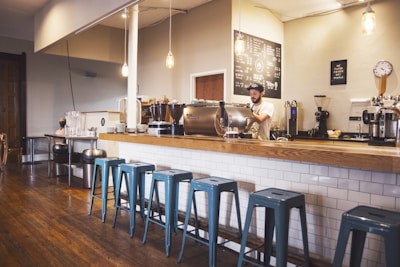 A cozy coffee shop interior featuring a wooden counter with a row of blue metal stools. A barista is preparing drinks behind the counter, surrounded by various coffee machines and accessories. Hanging light fixtures provide a warm ambiance, and a chalkboard menu is visible on the wall.