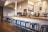 A cozy coffee shop interior featuring a wooden counter with a row of blue metal stools. A barista is preparing drinks behind the counter, surrounded by various coffee machines and accessories. Hanging light fixtures provide a warm ambiance, and a chalkboard menu is visible on the wall.