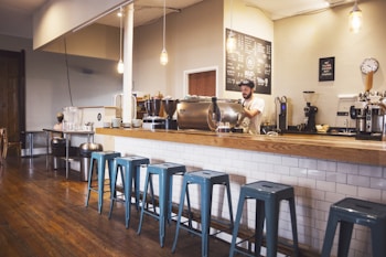 A cozy coffee shop interior featuring a wooden counter with a row of blue metal stools. A barista is preparing drinks behind the counter, surrounded by various coffee machines and accessories. Hanging light fixtures provide a warm ambiance, and a chalkboard menu is visible on the wall.