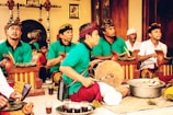 A group of men wearing traditional attire, including green shirts and batik sarongs, are seated and appear to be playing musical instruments as part of a cultural performance. They are surrounded by wooden structures that resemble gamelan instruments, with a gong visible in the background. The setting appears to be indoors with some traditional decor.