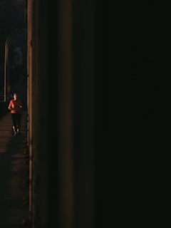 A person in a bright orange shirt is jogging on a path, partially hidden by darkness. The light contrasts sharply with the shadows that dominate the right side of the image.