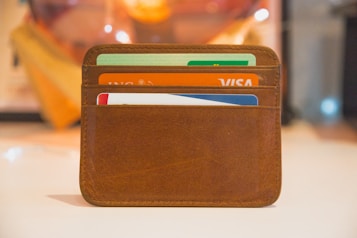 A brown leather wallet is holding several credit and payment cards. The wallet is neatly sewn and sits on a light surface with a warm, blurred background. The cards are of various colors, prominently featuring names of financial services.
