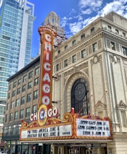 A vibrant marquee with the word 'CHICAGO' in large letters is displayed on a theater sign. The sign features decorative elements and lists upcoming shows. The building showcases ornate architectural details and is surrounded by modern skyscrapers against a bright blue sky with scattered clouds.