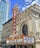 A vibrant marquee with the word 'CHICAGO' in large letters is displayed on a theater sign. The sign features decorative elements and lists upcoming shows. The building showcases ornate architectural details and is surrounded by modern skyscrapers against a bright blue sky with scattered clouds.