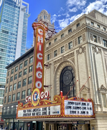 A vibrant marquee with the word 'CHICAGO' in large letters is displayed on a theater sign. The sign features decorative elements and lists upcoming shows. The building showcases ornate architectural details and is surrounded by modern skyscrapers against a bright blue sky with scattered clouds.