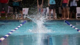 Water splashes dramatically at the center of a swimming pool, framed by lane dividers. In the background, a group of people stand by the edge of the pool, some with their legs visible. They appear to be spectators watching an event.