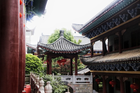 A traditional Asian architectural setting featuring ornate roofs with curved eaves and vibrant colors. Red lanterns are hanging, adding a touch of cultural significance. The scene is surrounded by lush greenery, and there are intricately carved pillars and stone railings.