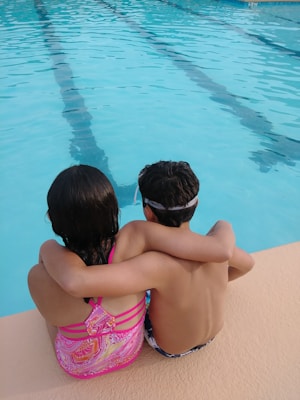 Two children are sitting together on the edge of a swimming pool, with their arms around each other. They are looking towards the water, which is clear and blue with lane markings visible. The child on the left is wearing a colorful pink swimsuit, while the child on the right is in swim trunks and a swim cap.