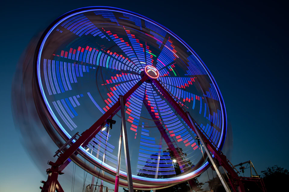 The image depicts a large, brightly lit Ferris wheel at night. The Ferris wheel is adorned with blue and red lights that create a mesmerizing pattern against the dark sky. The perspective of the photo is from below, looking up towards the towering structure. In the background, there are trees visible in the distance, adding depth to the scene.