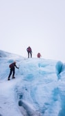 Several climbers are ascending an ice-covered slope, using ropes and climbing gear. The environment is stark and chilly, with expansive vistas of ice stretching into the horizon.