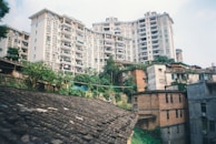 A cluster of tall, modern apartment buildings rises behind older, rustic brick structures with tiled roofs. Vegetation and trees are scattered around, indicating an urban setting transitioning into more traditional architecture.