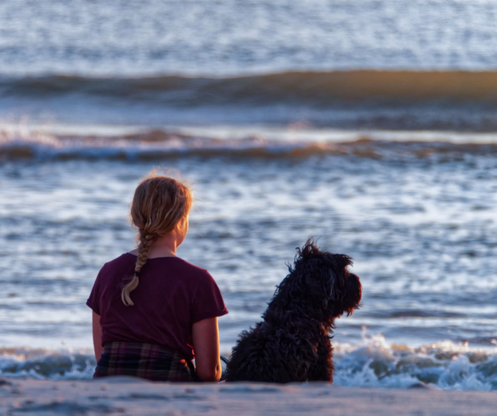 girl and dog sitting on seashore while watching on sea