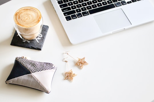 A neatly arranged workspace features a laptop with a white keyboard. Nearby, a glass of coffee sits on a slate coaster, emitting a creamy froth. A patterned clutch purse is placed beside star-shaped earrings crafted from a golden material.