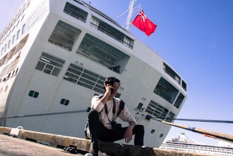 A person is sitting on a dock in front of a large white cruise ship. The ship is anchored, and a red flag is visible at the top. The individual is using a cellphone and has a camera around their neck. The sky above is clear and blue.