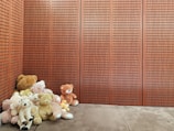A collection of teddy bears of various sizes and colors is arranged in the corner of a room with wooden paneled walls. The plush toys range in shades from brown to white and are huddled together on a soft, brown floor.