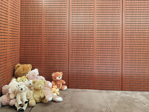 A collection of teddy bears of various sizes and colors is arranged in the corner of a room with wooden paneled walls. The plush toys range in shades from brown to white and are huddled together on a soft, brown floor.
