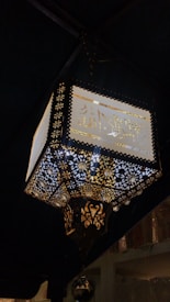 An intricately designed lantern with decorative Arabic calligraphy and ornate golden patterns. It is illuminated, casting light through its detailed cut-out designs, creating a warm and inviting ambiance.