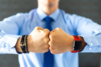 A person is wearing a blue dress shirt with their fists pressed together. A red smartwatch is on the left wrist, and several bracelets are on the right wrist. The background is blurred and dark gray.