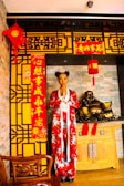A person is standing in traditional attire with a red floral design in a setting decorated for Chinese New Year. The backdrop includes intricate yellow and black patterned panels, red lanterns, and banners with Chinese characters. A wooden table holds a golden laughing Buddha statue, conveying a cultural and festive atmosphere.