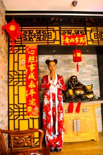 A person is standing in traditional attire with a red floral design in a setting decorated for Chinese New Year. The backdrop includes intricate yellow and black patterned panels, red lanterns, and banners with Chinese characters. A wooden table holds a golden laughing Buddha statue, conveying a cultural and festive atmosphere.