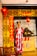 A person is standing in traditional attire with a red floral design in a setting decorated for Chinese New Year. The backdrop includes intricate yellow and black patterned panels, red lanterns, and banners with Chinese characters. A wooden table holds a golden laughing Buddha statue, conveying a cultural and festive atmosphere.