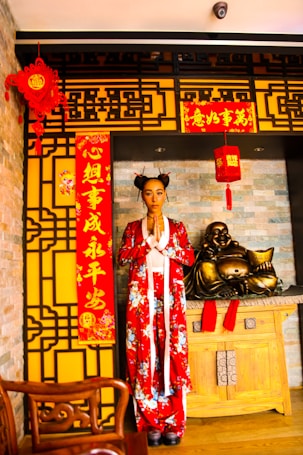 A person is standing in traditional attire with a red floral design in a setting decorated for Chinese New Year. The backdrop includes intricate yellow and black patterned panels, red lanterns, and banners with Chinese characters. A wooden table holds a golden laughing Buddha statue, conveying a cultural and festive atmosphere.