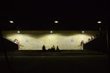 A dimly lit underground passage with concrete walls and illuminated by several lights. People are sitting or standing around, some on the stairs and platforms leading to the passage. A few individuals are gathered in small groups, creating a sense of community or waiting. The atmosphere appears subdued, with shadows adding to the ambiance.