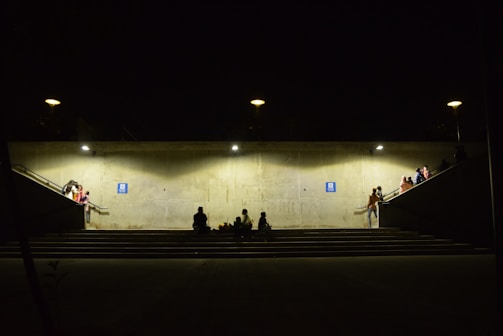 A dimly lit underground passage with concrete walls and illuminated by several lights. People are sitting or standing around, some on the stairs and platforms leading to the passage. A few individuals are gathered in small groups, creating a sense of community or waiting. The atmosphere appears subdued, with shadows adding to the ambiance.