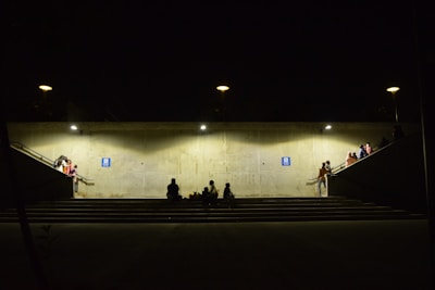 A dimly lit underground passage with concrete walls and illuminated by several lights. People are sitting or standing around, some on the stairs and platforms leading to the passage. A few individuals are gathered in small groups, creating a sense of community or waiting. The atmosphere appears subdued, with shadows adding to the ambiance.