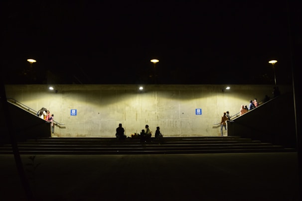 A dimly lit underground passage with concrete walls and illuminated by several lights. People are sitting or standing around, some on the stairs and platforms leading to the passage. A few individuals are gathered in small groups, creating a sense of community or waiting. The atmosphere appears subdued, with shadows adding to the ambiance.
