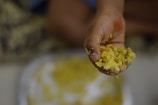 A close-up of a hand holding a portion of rice, mixed with small pieces of vegetables, likely a homemade dish. The background shows a blurred plate with more of the same food.