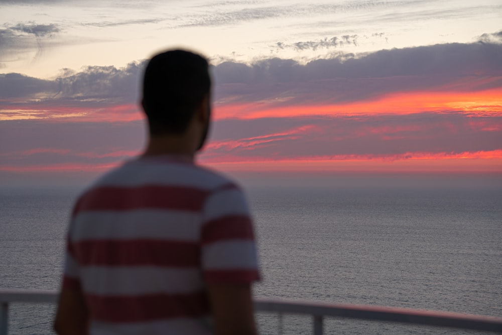 man wearing white and red striped shirt standing near the body of water during golden hour