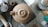 A clay teapot is centered on a workbench surrounded by various pottery tools and materials. The surface around the teapot is scattered with other pottery-related objects, such as cups and brushes. The floor appears to be concrete, and there are a pair of shoes visible at the bottom right corner of the image.