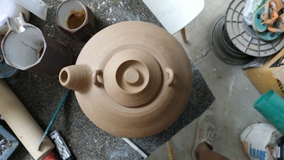 A clay teapot is centered on a workbench surrounded by various pottery tools and materials. The surface around the teapot is scattered with other pottery-related objects, such as cups and brushes. The floor appears to be concrete, and there are a pair of shoes visible at the bottom right corner of the image.