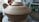 A large, unglazed ceramic pot with a lid, situated on a pottery wheel. The setting appears to be a pottery studio with various tools and materials visible in the background.