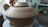 A large, unglazed ceramic pot with a lid, situated on a pottery wheel. The setting appears to be a pottery studio with various tools and materials visible in the background.