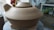 A large, unglazed ceramic pot with a lid, situated on a pottery wheel. The setting appears to be a pottery studio with various tools and materials visible in the background.
