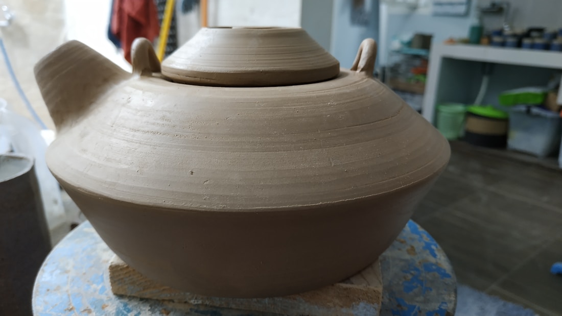 A large, unglazed ceramic pot with a lid, situated on a pottery wheel. The setting appears to be a pottery studio with various tools and materials visible in the background.