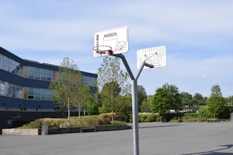A basketball hoop stands in an empty outdoor court surrounded by trees and a modern building with many windows. The area appears clean and well-maintained, with a clear blue sky above.