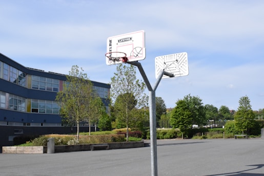 A basketball hoop stands in an empty outdoor court surrounded by trees and a modern building with many windows. The area appears clean and well-maintained, with a clear blue sky above.