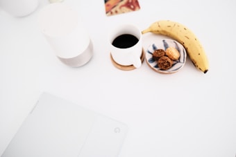 A minimalist setup with a white table featuring a banana, a cup of black coffee, a small plate with cookies, and a white electronic device. The ambiance is clean and organized, with a focus on simplicity.