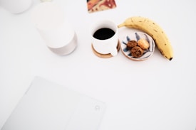 A minimalist setup with a white table featuring a banana, a cup of black coffee, a small plate with cookies, and a white electronic device. The ambiance is clean and organized, with a focus on simplicity.