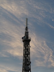A tall communication tower stands against a backdrop of soft, wispy clouds in a sky tinged with shades of blue and gray. The structure is equipped with various antennas and satellite dishes.