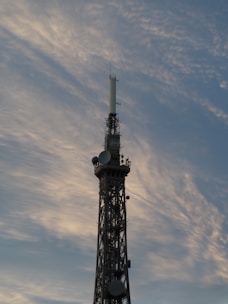 A tall communication tower stands against a backdrop of soft, wispy clouds in a sky tinged with shades of blue and gray. The structure is equipped with various antennas and satellite dishes.
