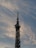 A tall communication tower stands against a backdrop of soft, wispy clouds in a sky tinged with shades of blue and gray. The structure is equipped with various antennas and satellite dishes.