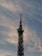 A tall communication tower stands against a backdrop of soft, wispy clouds in a sky tinged with shades of blue and gray. The structure is equipped with various antennas and satellite dishes.