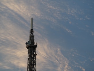 A tall metal communication tower extends into the sky, featuring various antennas and satellite dishes. The backdrop is a sky filled with soft, wispy clouds that catch the golden hues of the setting sun.
